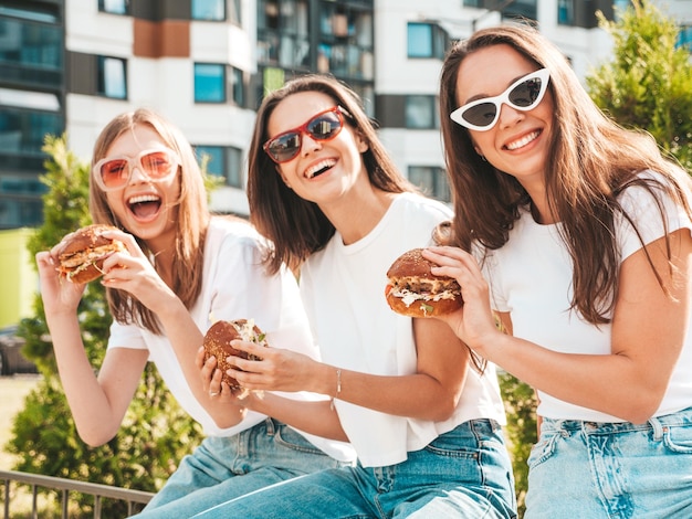 トレンディな夏の同じ服を着た3人の若い美しい笑顔のヒップスター女性ストリートでポーズをとるセクシーな屈託のない女性サングラスで楽しんでいるポジティブモデルジューシーなハンバーガーを持ってハンバーガーを食べる