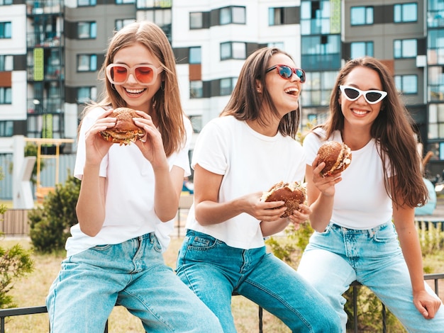 Three young beautiful smiling hipster female in trendy summer\
same clothes sexy carefree women posing in the streetpositive\
models having fun in sunglassesholding juicy burger and eating\
hamburger