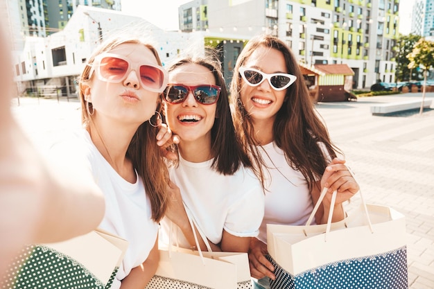 Three young beautiful smiling hipster female in trendy summer same clothes Sexy carefree women posing in the streetPositive models having fun in sunglassesHolding shopping bagsTaking Pov selfie