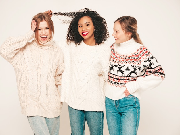 Three young beautiful smiling hipster girls in trendy winter sweaters. Positive models having fun and hugging