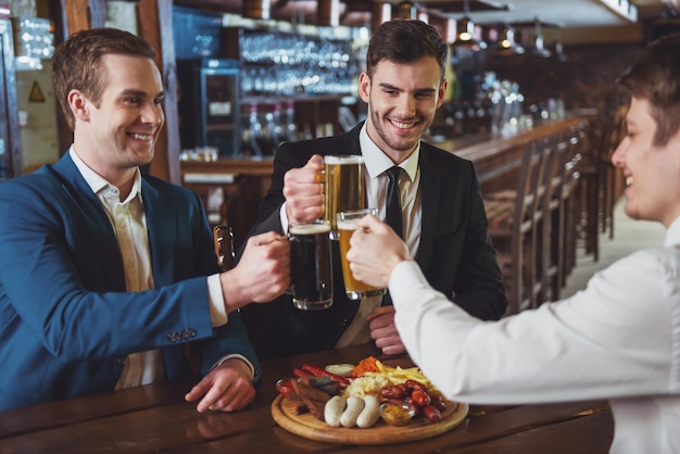 Three young businessmen in suits are smiling and clanging glasses of beer together while sitting in pub