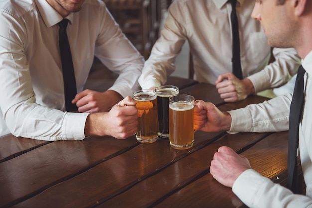 흰색 클래식 셔츠를 입은 세 명의 젊은 사업가들이 펍 클로즈업에 앉아 있는 동안 맥주 잔을 들고 있다