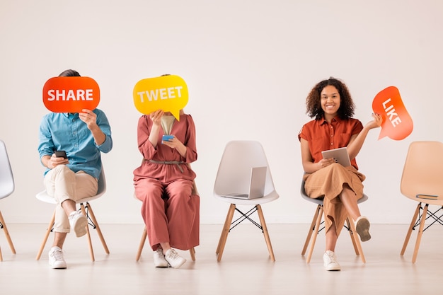 Three young casual friends with mobile gadgets holding multi-color paper speech bubbles while communicating in social networks