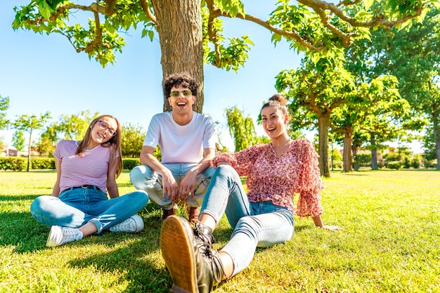 写真 肖像画のためにカメラを見て微笑んで緑の公園のフィールドで休んでいる3人の若い多様な友人。自然の中で時間を過ごし、都市生活の日常を破るz世代の学生。友情は何の違いもありません