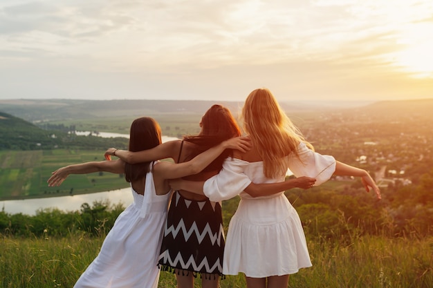 Three young female friends is hugging and having summer picnic on a hill at sunset.