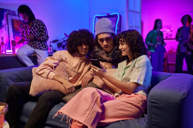 Three young friends in casualwear watching online videos in smartphone of girl