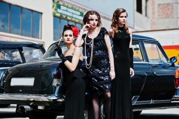 Foto tre giovani ragazze in abito in stile retrò vicino vecchie auto d'epoca classiche.