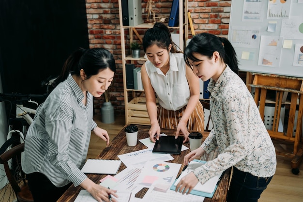 three young girls coworkers tidy documents after meeting in board room and leaving. female colleagues at table standing finding papers. creative team partner girls in small business company.