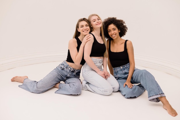 Three young interracial women in black tops and jeans look at camera sitting on their knees against white background