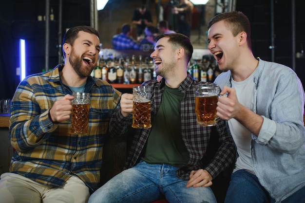 Three young men in casual clothes are smiling and clanging glasses of beer together while sitting at bar counter in pub