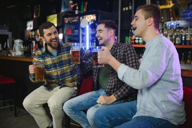 カジュアルな服を着た 3 人の若い男性が、パブのバーカウンターに座って、ビールのグラスを一緒にカチャカチャ鳴らしながら笑っている