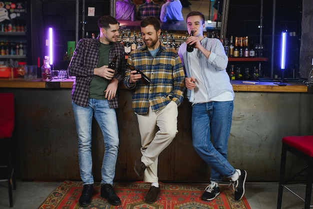 Three young men in casual clothes are smiling holding bottles of beer while standing near bar counter in pub