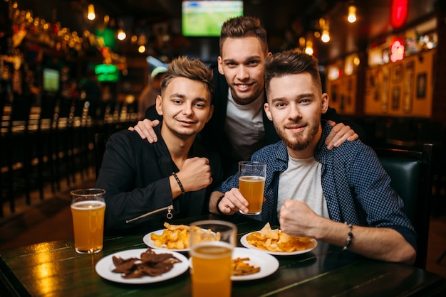 Three young men poses at the table with beer, crisps and crackers, sport bar interior, happy friendship of football fans