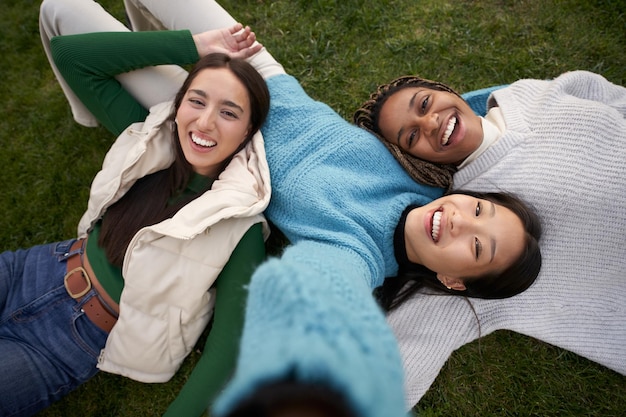 Foto tre giovani ragazze multirazziali sdraiate sull'erba che fanno un selfie con il cellulare guardando la telecamera