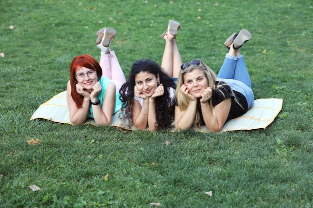 Photo three young smiling girlfriends lie on blanket on the green grass and pose for the camera