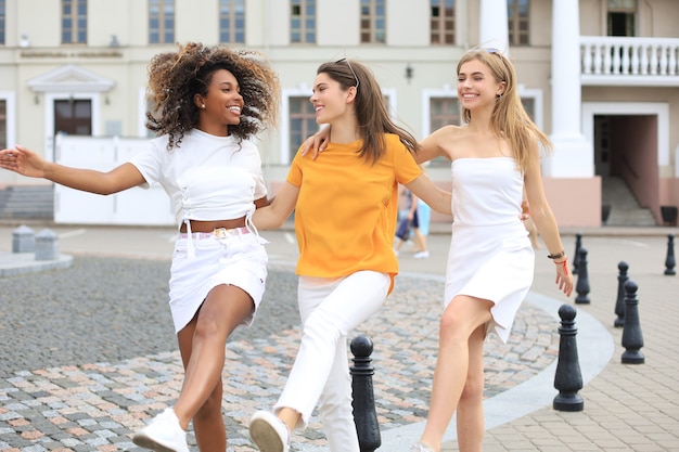 Three young smiling hipster women in summer clothes posing on street.Female showing positive face emotions. Dancing.