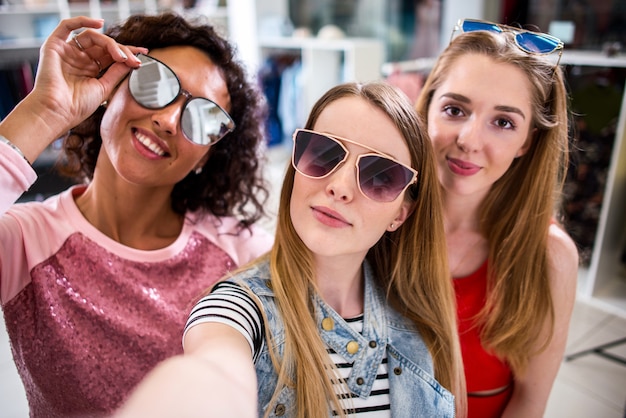 Tre giovani amiche alla moda che sollevano gli occhiali da sole alla moda mentre prendono il selfie