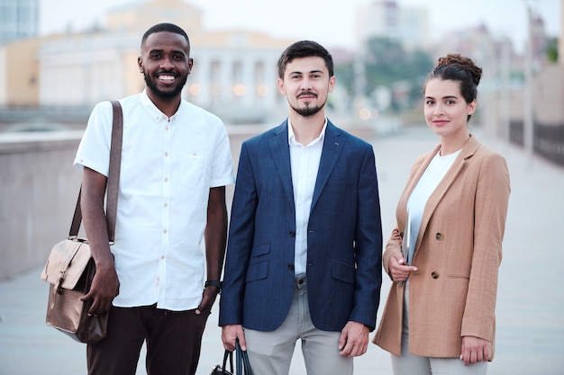 Three young successful intercultural business partners in smart casualwear standing in row in front of camera against urban environment