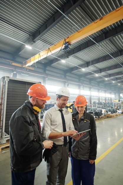 Three young workers of industrial plant discussing technical sketch of machine detail at meeting in workshop