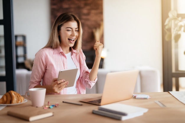 Thrilled young lady rising fist victoriously and holding tablet while sitting at desk with laptop on it