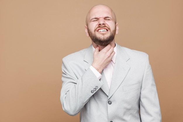 Throat pain or cold. portrait of middle aged bald bearded businessman with closed eyes in light gray suit standing and touching his painful neck. indoor studio shot, isolated on light brown background