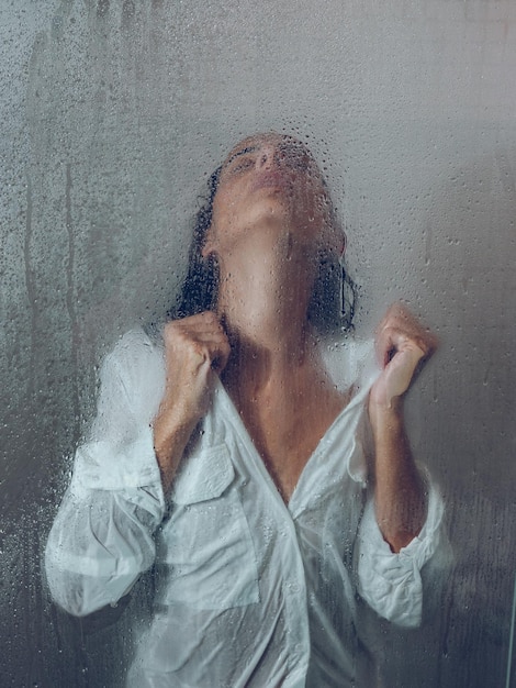 Foto attraverso il vetro di una donna attraente con una camicetta bagnata in piedi in una cabina doccia leggera con porta trasparente e gocce d'acqua nel bagno