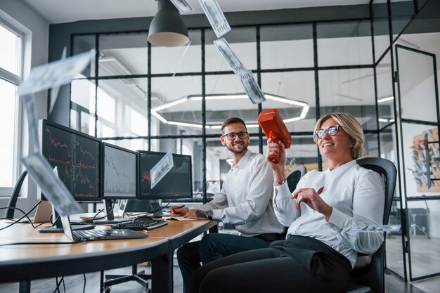 Throwing money to the air by special device. Two stockbrokers in formal clothes have a break and fun in the office.