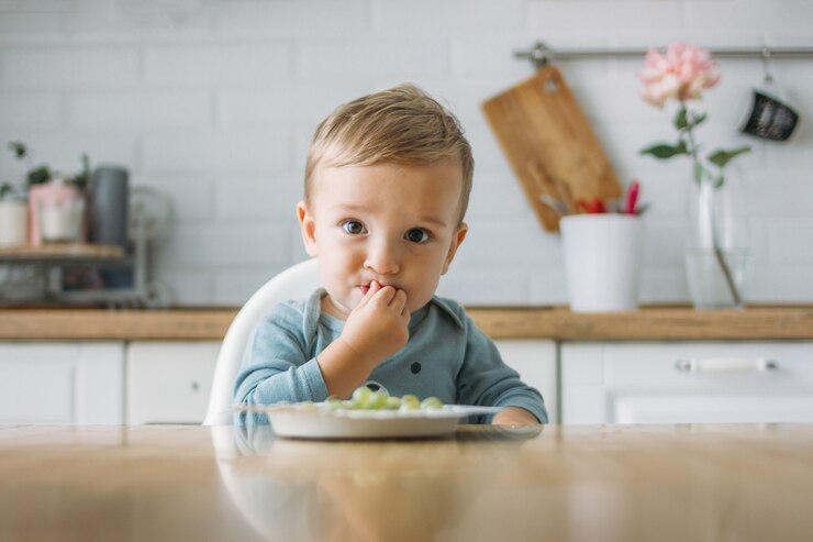 Thuis charmerend weinig babyjongen die eerste voedsel groene druif eten bij heldere keuken