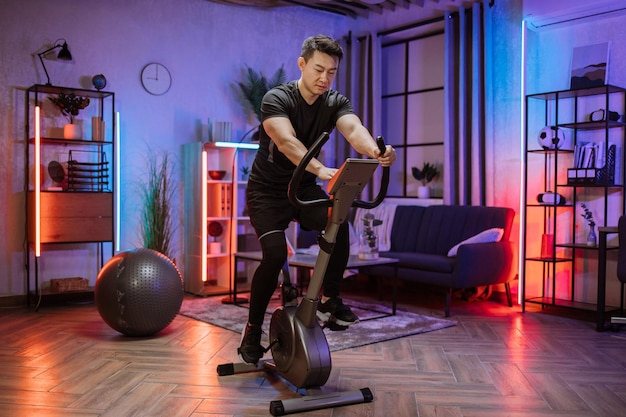 Thuis fitness training jonge aziatische man atleet training op slimme stationaire fiets binnenshuis