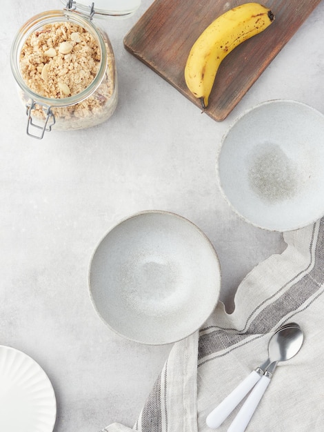 Thuis gedekte tafel voorbereid voor ontbijt of snack grijze kommen lepels banaan op houten plank en glazen pot met muesli bovenaanzicht met kopieerruimte