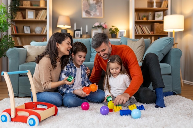 Thuis geniet een gezin van vier personen van hun vrije tijd samen. Kinderen spelen met speelgoed terwijl ouders zich bij hen voegen op de vloer. Lachen weerklinkt in hun gezellige ruimte.