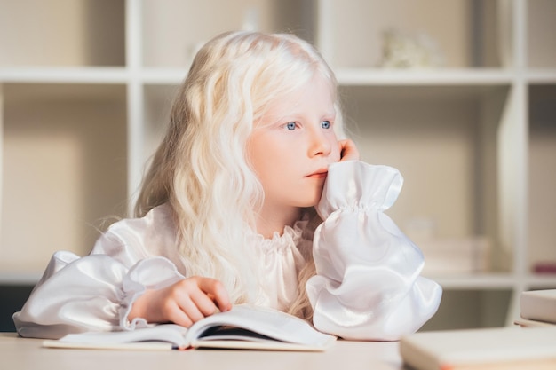 Thuis leren. Zelfisolatie van kinderen. Eenzaamheid in quarantaine. Verveeld doordacht schattig blond albino-meisje in witte blouse zittend met open boek opzij kijkend.