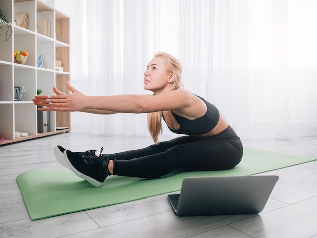 Thuis stretching sportieve vrouw genieten van fitness Online les atletische dame zwarte sportkleding opwarming van de aarde buigen voorwaartse training op laptop training in lichte kamer interieur doen