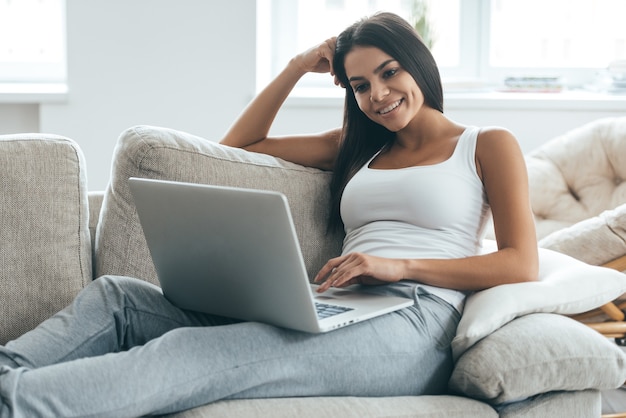 Thuis surfen op internet. Aantrekkelijke jonge vrouw die op laptop werkt en glimlacht terwijl ze thuis op de bank ontspant