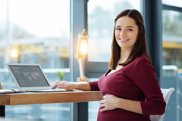 Thuis uitrusten. Close-up van lachende zwangere vrouw met behulp van een laptop terwijl ze haar buik aanraakt en optimisme uiten