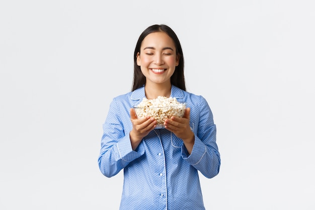 Thuis vrije tijd, logeerpartij en slaapfeestje concept. Glimlachend tevreden Aziatisch meisje genieten van vrije dag in bed met popcorn, eten en films kijken in pyjama's, staande witte muur.