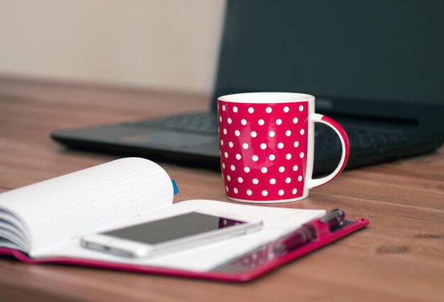 Thuis werken - computer en notitieboekje met theekopje. Kopje cappuccino en notebook in de buurt van telefoon en laptop op houten tafel.