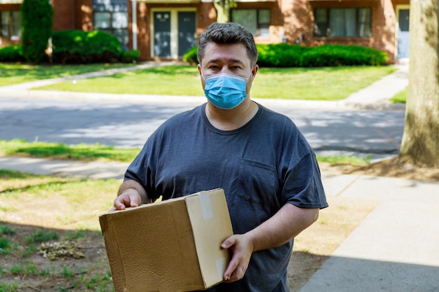 Thuisbezorging, online bestellen van een man in een medisch masker met een doos, een pakket in zijn handen voedselbezorging tijdens de quarantaine van de coronaviruspandemie.