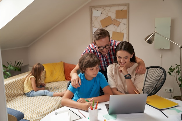 Thuisonderwijs jonge positieve ouders die zijn zoon helpen met huiswerk terwijl een klein schattig meisje zit