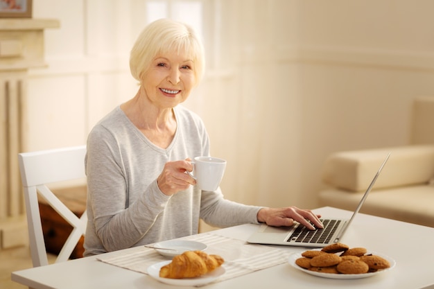 Thuiswerken. Mooie blonde die een glimlach op haar gezicht houdt en de vingers op het toetsenbord legt terwijl ze thee gaat drinken