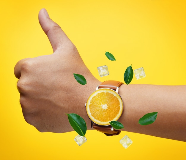 フルーツオレンジ色の腕時計を身に着けている親指の手を緑の葉とアイスキューブが飛ぶ
