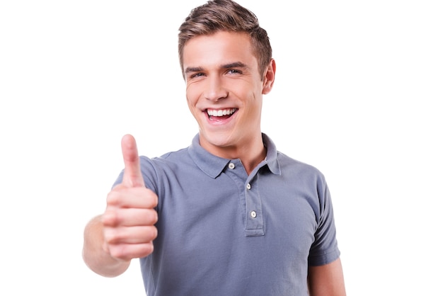 Thumb up! Happy young man looking at camera and showing his thumb up while standing isolated on white background