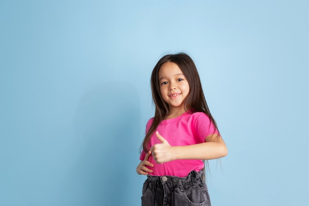 Thumb up, nice gesture. Caucasian little girl's portrait on blue  wall. Beautiful female model in pink shirt. Concept of human emotions, facial expression, youth, childhood.