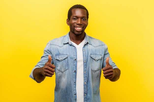 Thumbs up, excellent job. Portrait of enthusiastic handsome man in denim casual shirt with rolled up sleeves smiling and showing like gesture at camera. studio shot isolated on yellow background