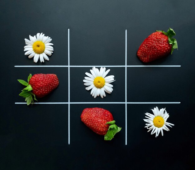 Tic tac toe op een zwarte achtergrond gemaakt van kamille bloemen en aardbeien. Plat leggen. Natuurlijk begrip.