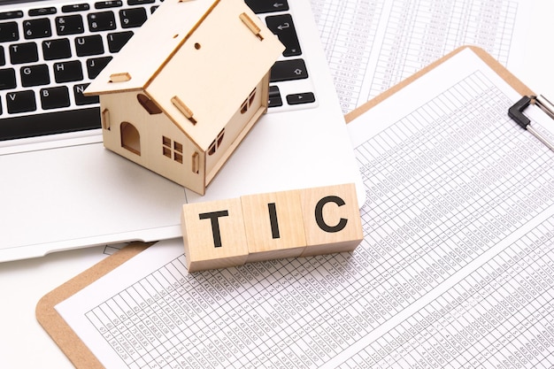 Слово TIC на деревянных кубиках на белом фоне с клавиатурой настольного ноутбука и небольшим деревянным домом