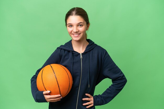 Tiener blank meisje speelt basketbal over geïsoleerde achtergrond poseren met armen op heup en glimlachen