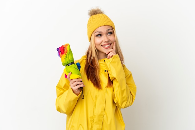 Tiener blond meisje draagt een regenbestendige jas over een geïsoleerde witte achtergrond en denkt aan een idee terwijl ze omhoog kijkt