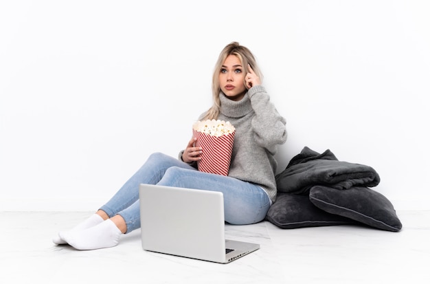 Tiener blond meisje eet popcorn tijdens het kijken naar een film op de laptop met twijfels en denken