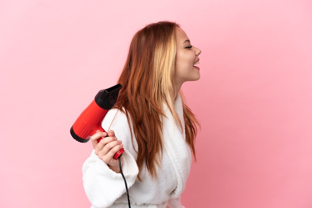 Tiener blond meisje met een haardroger over een geïsoleerde roze achtergrond die lacht in zijpositie